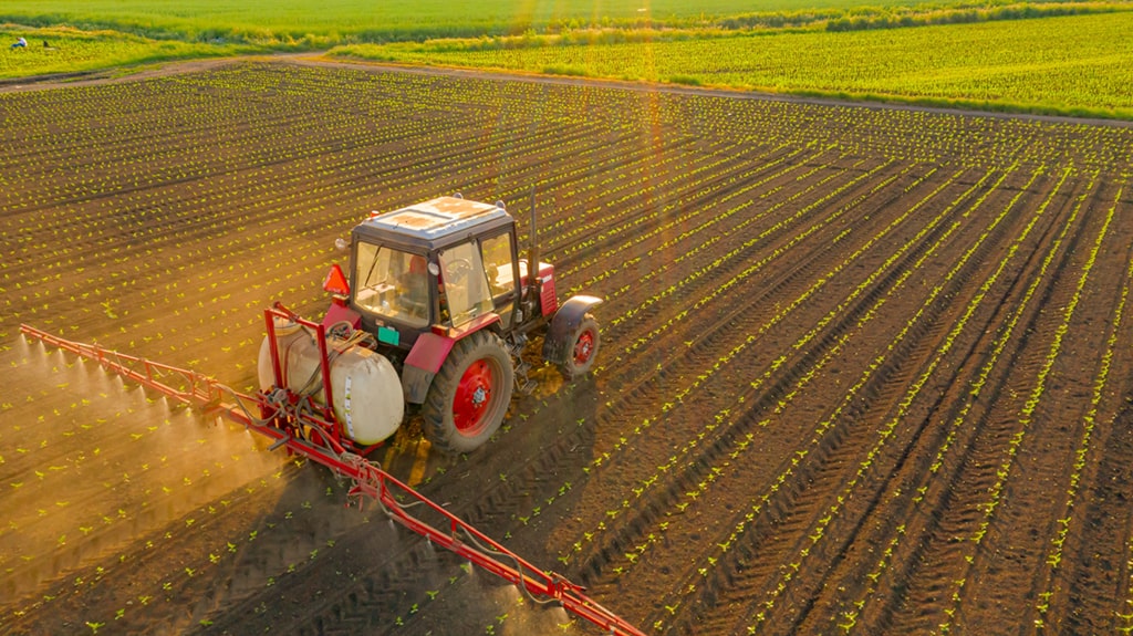 A red tractor spraying crops in a large agricultural field during sunset.